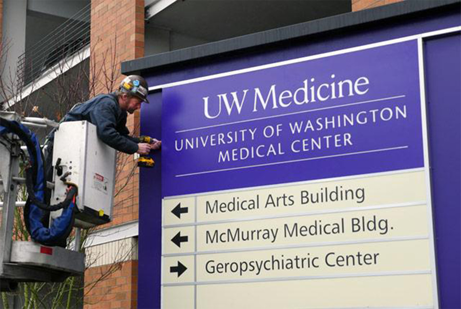 A photo of a worker installing a new purple directional sign on the UWMC – Northwest Campus. The sign says Medical Arts Building left, McMurray Medical Building (left), and Geropsychiatric Center (right). A photo of a worker installing a new purple directional sign on the UWMC – Northwest Campus. The sign says Medical Arts Building left, McMurray Medical Building (left), and Geropsychiatric Center (right).